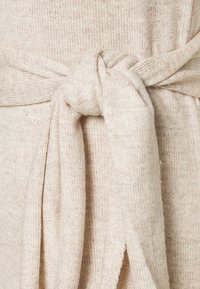 Maglione beige lavorato a maglia con dettaglio di cintura annodata in vita, caratterizzato da un tessuto morbido e testurizzato e un leggero motivo a macchie.