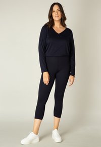 Marineblauw shirt met lange mouwen en een V-hals, gecombineerd met bijpassende cropped leggings, beide gemaakt van een soepele, rekbare stof, gedragen met witte platform sneakers.