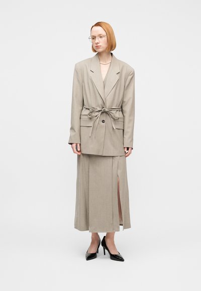 Γυναίκα με κοντά κόκκινα μαλλιά, φοράει μπεζ σακάκι με ζώνη, μακριά φούστα με σκίσιμο, γυαλιά, κολιέ με πέρλες και μαύρες ψηλοτάκουνες γόβες, στέκεται ακίνητη.