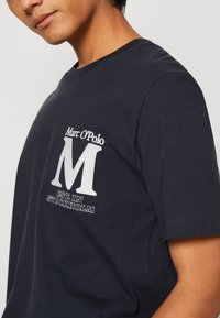 Granatowa bawełniana koszulka z białym haftowanym logo oraz napisem "Marc O'Polo" i "EST. W STOCKHOLM" z przodu. Klasyczny krój z okrągłym dekoltem.
