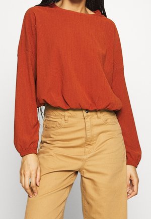 Vrouw draagt een roestkleurige blouse met lange mouwen, ingestopt in een beige broek met een hoge taille, tegen een lichte achtergrond.