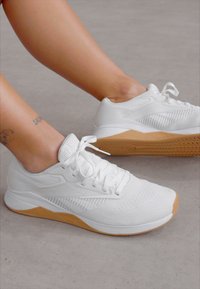 Λευκά αθλητικά παπούτσια με υφή πλέγματος στην άνω πλευρά και καουτσούκ σόλα. Τα χαρακτηριστικά περιλαμβάνουν κορδόνια, επενδεδυμένο κολάρο και ανοιχτό καφέ πάτο.