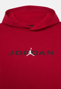 Červená mikina s kapucňou vyrobená z mäkkej látky, obsahujúca čierne logo "JORDAN" a bielu siluetu, s klokaním vreckom a nastaviteľnou kapucňou.