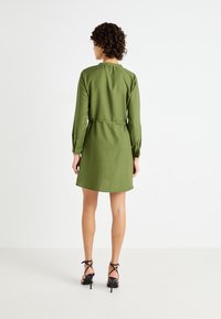 Robe verte à manches longues avec une taille cintrée et une coupe droite. Le tissu a une texture lisse et l'encolure est ronde. Talons noirs à lanières.