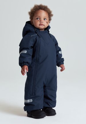 Småbarn med krøllet hår iført en mørkeblå vandtæt flyverdragt med reflekterende striber og sorte støvler, der står foran en ensfarvet baggrund.