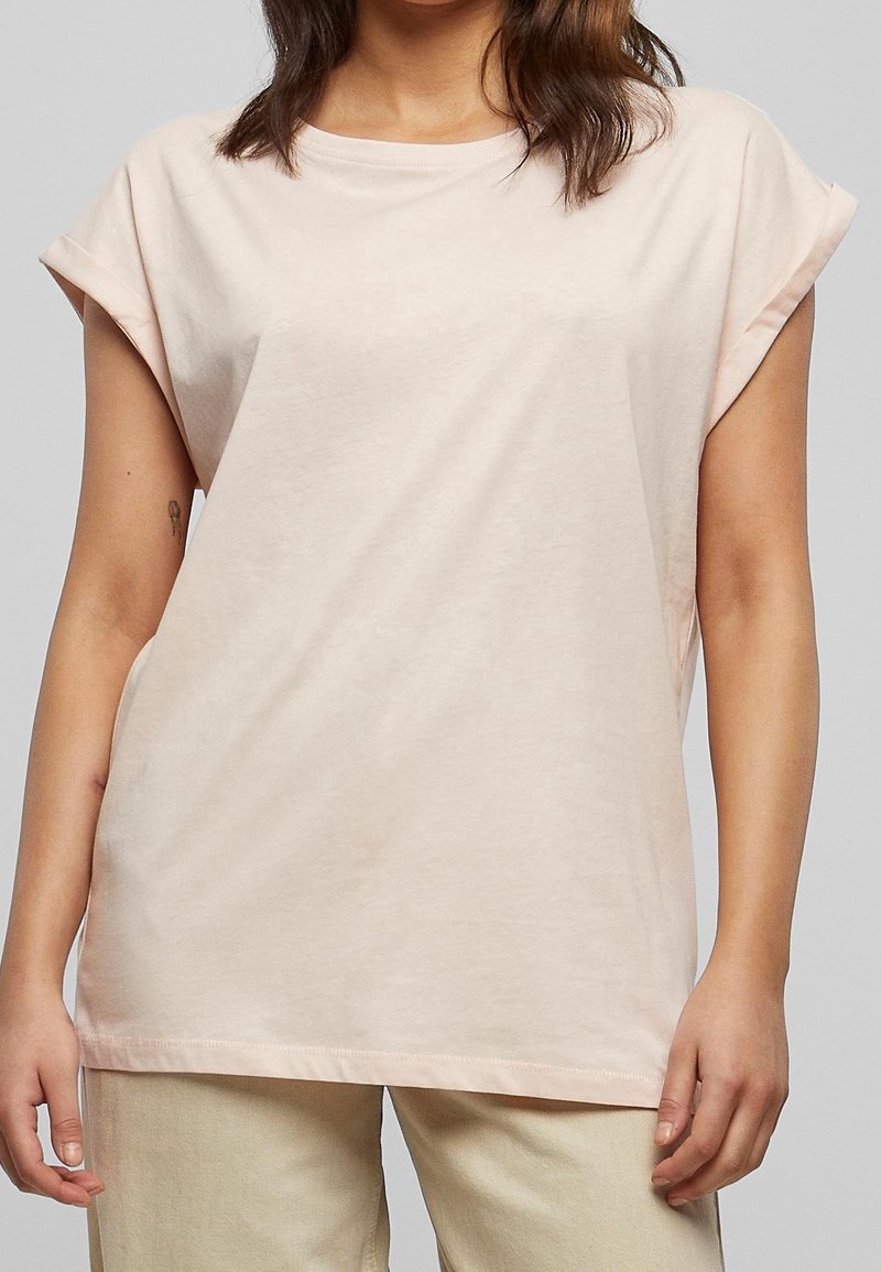 Femme portant un t-shirt rose clair à manches courtes et un pantalon beige, cadré des épaules jusqu'à mi-cuisse sur un fond gris uni.