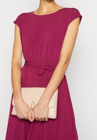 Fuksjowa sukienka z krótkimi rękawami i związywanym w talii paskiem, trzymająca beżową torebkę z złotymi akcentami. Gładki materiał i luźny krój.