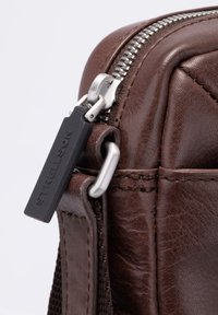 Dettaglio di borsa in pelle marrone con cerniera metallica, accessori in argento e superficie strutturata con una targhetta nera con la scritta "Strellson."