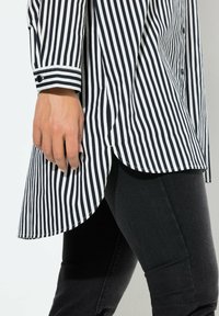 Maglia a righe bianche e nere con polsini abbottonati, caratterizzata da un orlo posteriore più lungo, abbinata a leggings neri. Tessuto liscio, vestibilità sagomata.