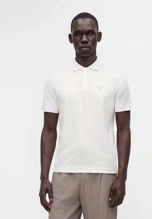 Homme portant un polo blanc uni à manches courtes et un pantalon beige clair, debout devant un fond blanc.