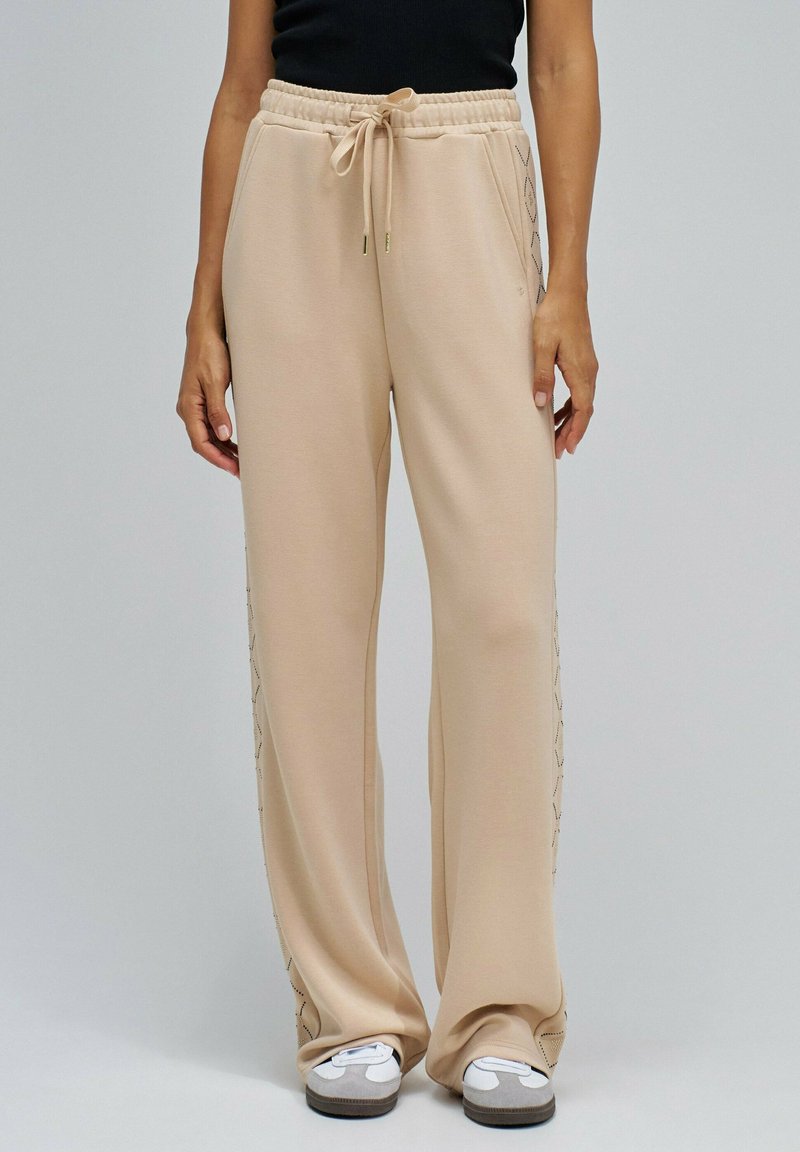 Beige joggers van zacht materiaal, met een trekkoord in de taille, zijzakken en een decoratief patroon langs de buitenste naden.