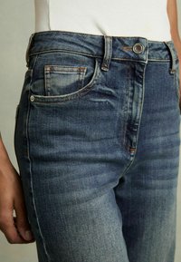 Jean en denim noir avec des dégradés clairs, comportant cinq poches, des accents cousus et une fermeture à bouton en métal à la taille.