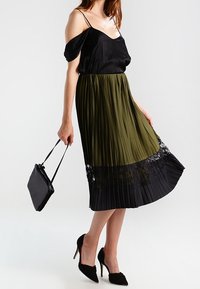 Top in satin nero a spalle scoperte, gonna plissettata oliva con bordo in pizzo nero, abbinata a una borsa nera in vernice e tacchi a punta.