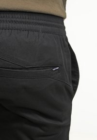 Pantalons cargo noirs extensibles avec taille élastique, présentant une poche arrière avec une petite étiquette blanche "VOLCOM". Texture de tissu lisse.