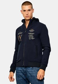 Felpa con zip color navy realizzata in tessuto morbido, con dettagli del logo e del testo ricamati, due tasche laterali e polsini e orlo a costine.