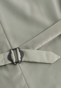 Tela de color gris claro con una textura suave, que presenta una hebilla de metal negro y correas a juego. El diseño destaca por sus líneas y ángulos entallados.