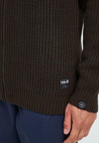 Brauner, gestrickter Zip-Cardigan mit gerippter Textur, Logo-Patches und kontrastierendem Stoff an Kragen und Bündchen.