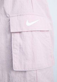 Materialul jachetei roz deschis are o finisare texturată, cu o buzunare mare pe mâneca stângă și un logo alb Nike pe clapeta buzunarului.