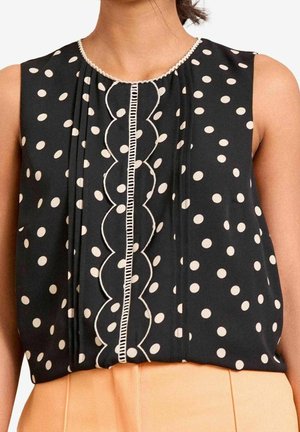 Mouwloze zwarte blouse met witte polka dots, gegolfde verticale afwerking, en beige broek gedragen door een persoon met een paardenstaart.