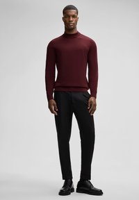 Een man staat zelfverzekerd in een strakke bordeauxrode coltrui, gecombineerd met zwarte broek en schoenen, voor een neutrale achtergrond.