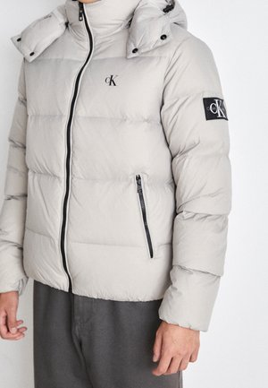 Lysegrå dunjakke med frontlynlås, lynlås på sidelommen og sorte Calvin Klein-logoer på brystet og venstre ærme.