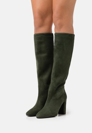Bottes mi-mollet en daim vert foncé avec des bouts pointus et des talons blocs portées sur des jambes nues sur fond blanc.