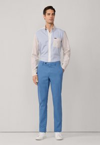 Lichtblauwe overhemdbloes met beige mouwen en een borstzak, gecombineerd met blauwe broeken en witte sneakers. Glad materiaal en op maat gemaakte pasvorm.