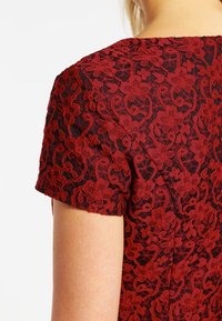 Dos et épaule d'une personne portant un haut noir à manches courtes avec un tissu en dentelle florale rouge détaillée en superposition.