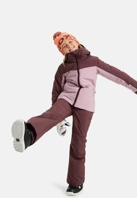 Skijack in donker bordeauxrood en lichtroze, gecombineerd met bijpassende broek. Oranje beanie met een pompon, sneeuwlaarzen en gebreide handschoenen.