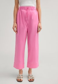 Roze wijde broek met een trekkoord in de taille en omgeslagen manchetten, gemaakt van gladde stof. G gedragen met beige slippers met zwarte letters.