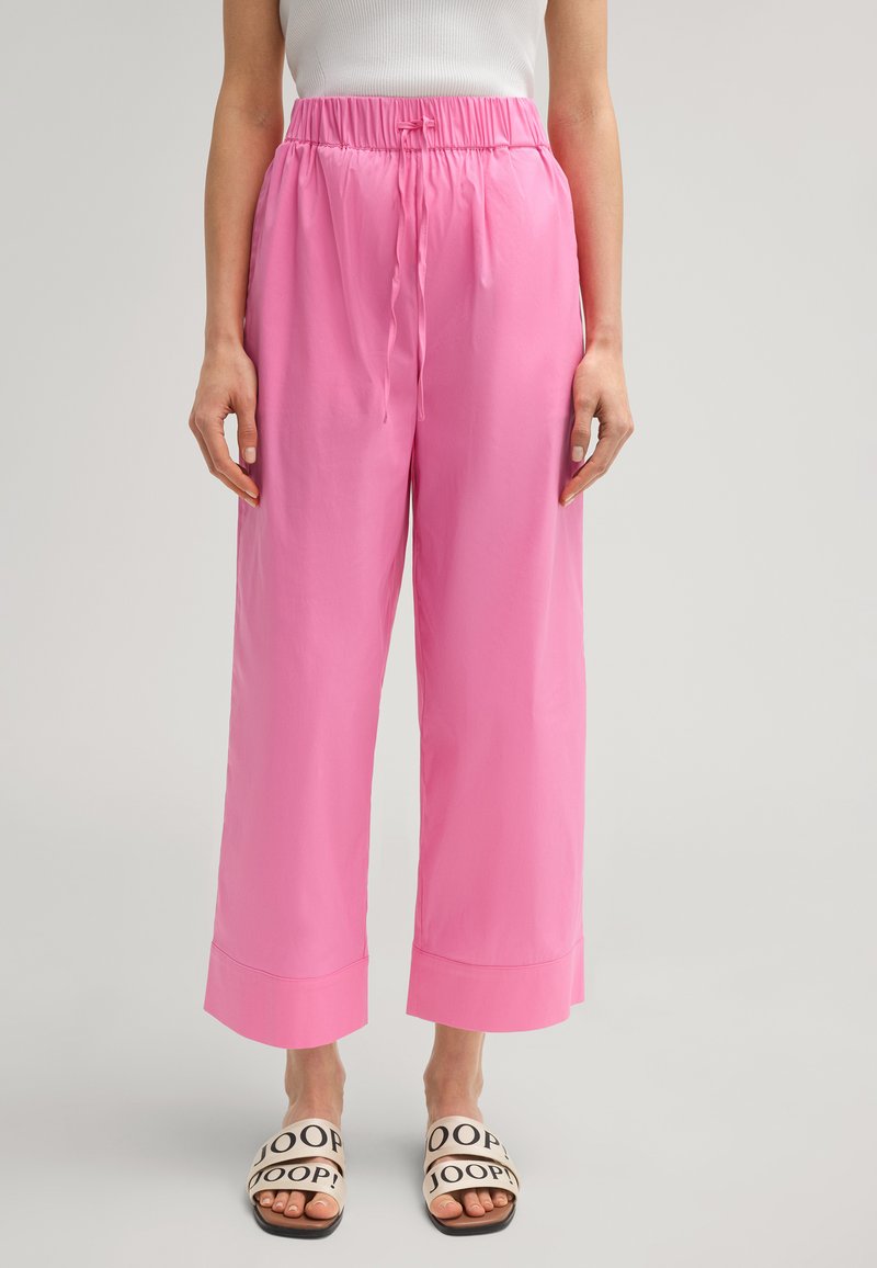Roze wijde broek met een trekkoord in de taille en omgeslagen manchetten, gemaakt van gladde stof. G gedragen met beige slippers met zwarte letters.