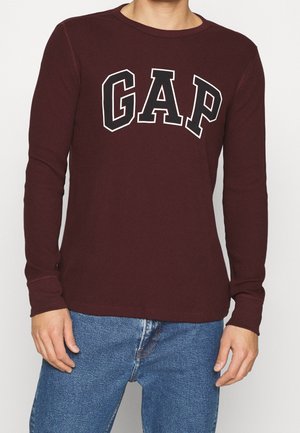 Man wearing a dark red long-sleeve shirt with white-outlined "GAP" lettering and blue jeans, standing against a plain background.