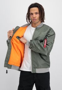 Jachetă bomber verde măsliniu cu guler și manșete ribbate gri, căptușeală portocalie, închidere cu fermoar și buzunar lateral. Detaliu cu etichetă roșie pe mânecă.