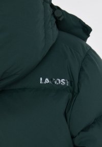 Tamnozeleni puffer kaput s kvadratnim uzorkom, sjajnim srebrnim logotipom "LACOSTE" i podesivom kapuljačom sa detaljem kopčanja.