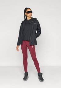 Čierna nepremokavá bunda s kapucňou, čierne tričko s krátkym rukávom, gaštanovočervené legíny a čierne športové topánky; modelka so slnečnými okuliarmi.