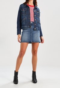 Chaqueta de mezclilla en azul oscuro con dos bolsillos en el pecho y botones de cobre, combinada con una falda de mezclilla azul clara. Botines negros completan el conjunto.