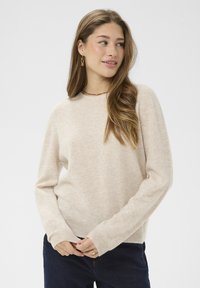 Beige hosszú ujjú pulóver, kerek nyakkivágással, puha textúrával, bordás mandzsettával és aljjal, sötét farmernadrággal párosítva.