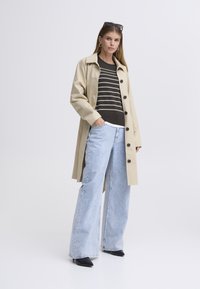 Trench beige avec de gros boutons porté sur un pull rayé foncé, associé à un jean large bleu clair et des chaussures noires à bout pointu.