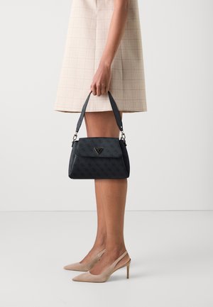 Mujer con vestido beige y tacones sosteniendo un bolso negro de estampado con asa superior contra un fondo sencillo.