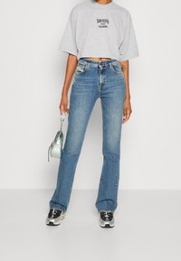 Camiseta gris corta con texto negro, jeans acampanados azul, zapatillas plateadas y negras, sosteniendo un pequeño bolso transparente con cadena.