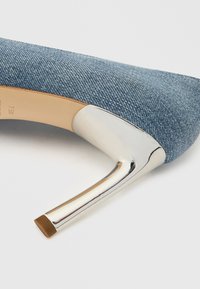 Pantof cu toc înalt din denim, cu un toc metalic argintiu lucios și vârf rotund, având o textură moale și un material albastru deschis.