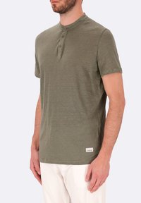 Maglietta di cotone verde con colletto corto e rotondo e tre bottoni frontali. Etichetta notevole sull'orlo. Il modello indossa pantaloni bianchi.