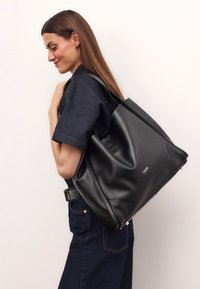 Bolso tote de cuero negro con un diseño suave y caído. Presenta un acento del logo dorado y asas resistentes, combinado con un conjunto de denim.