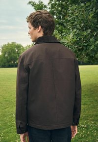 Chaqueta marrón con un cuello de pana, de corte recto y con puños abotonados. Bolsillos visibles a los lados, combinada con jeans oscuros.