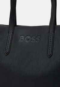 Czarna skórzana torba tote z wytłoczonym logo "BOSS" i dwiema przeszytymi uchwytami na jednolitym jasnym tle.