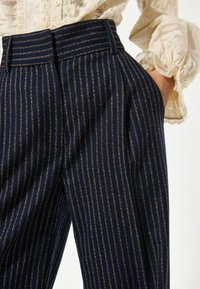 Pantaloni blu navy con strisce dorate verticali, caratterizzati da una vita alta, pieghe e una texture morbida. Primo piano sull'area della tasca.