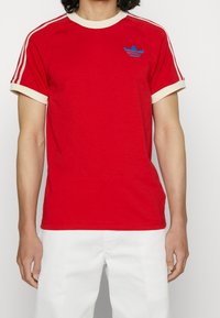 Červené tričko s krátkým rukávem s bílým obratlem a manžetami, vyšitým modrým logem Adidas na hrudi a kontrastními bílými postranními pruhy.