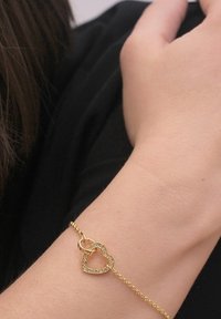 Bracciale in oro con pendente a forma di cuore e finitura scintillante. Il design presenta forme di cuori intrecciati. Texture liscia.