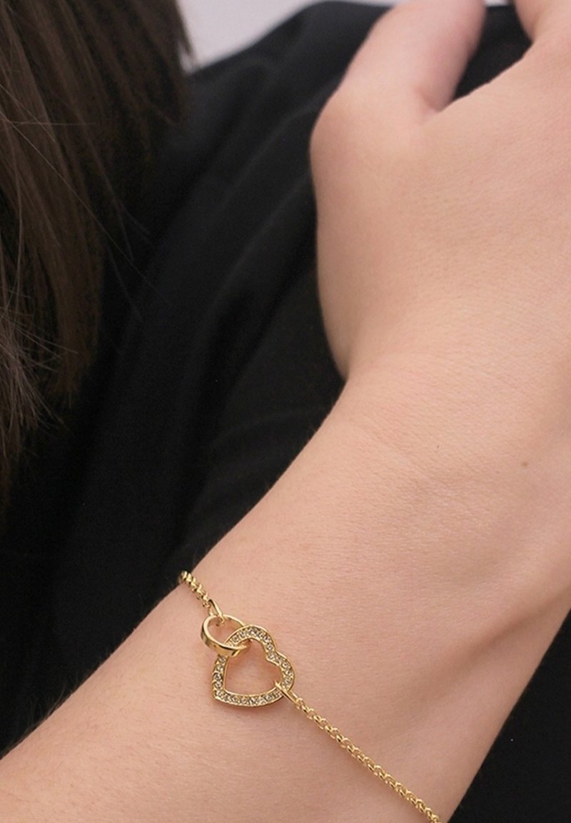 Bracciale in oro con pendente a forma di cuore e finitura scintillante. Il design presenta forme di cuori intrecciati. Texture liscia.