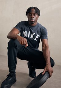 Pelēks Nike T-krekls ar balto logo, melni sporta bikses un melni apavi. Personas rokās ir melna svara plāksne, un viņa ar kustību ir piekususi.